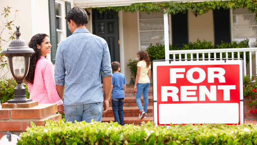 A woman smiles at a man while two children walk up the stairs to a house. A "for rent" sign is in the front yard.