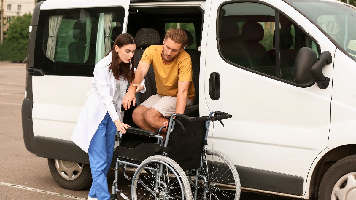 A female wearing scrubs and a doctor's coat helps a man out of a van and into a wheelchair.